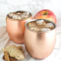 Two copper mugs of Ginger Apple Cider Punch with ice cubes floating in them and ginger, cinnamon, and apple garnishes on the side from acleanplate.com