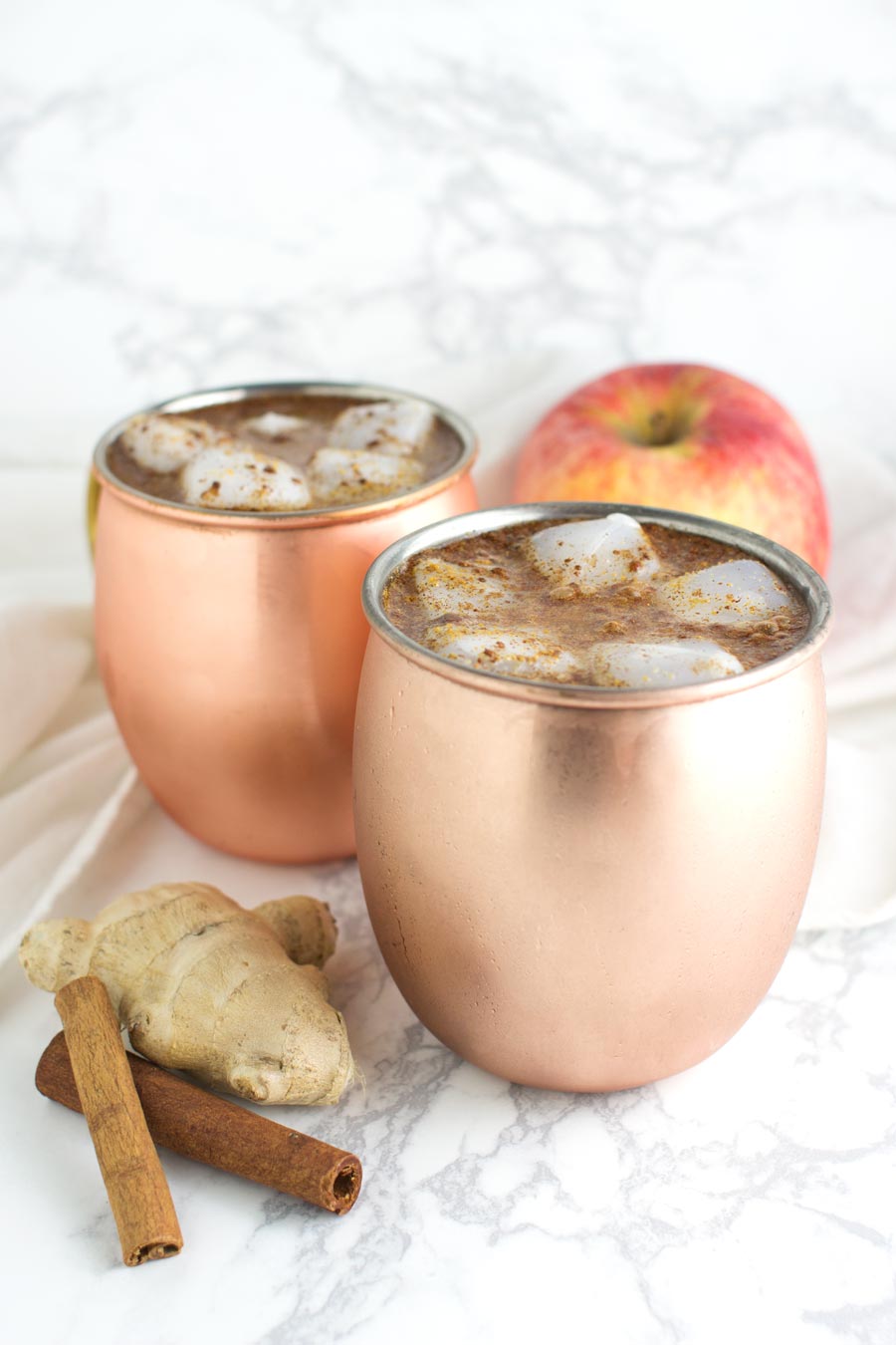 Two copper mugs of Ginger Apple Cider Punch with ice cubes floating in them and ginger, cinnamon, and apple garnishes on the side from acleanplate.com